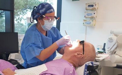 A person in a mask, safety glasses, gloves and hair net shines a light into the mouth of a prone man in pink clothing.